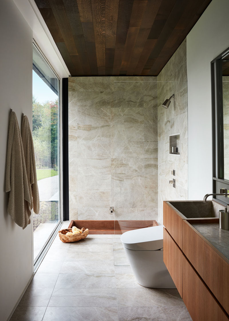 In this modern bathroom, a glass shower screen almost appears invisible, and the bath has been sunken down into the floor beside the shower, both of which have a view of the trees outside. #ModernBathroom #BathroomDesign #SunkenBathtub