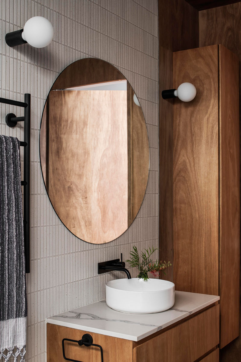 This modern bathroom features tile covered walls, a wood vanity and matching cabinetry, and a round mirror with sconces either side. #ModernBathroom #RoundMirror #WoodVanity #Sconces #TiledWall