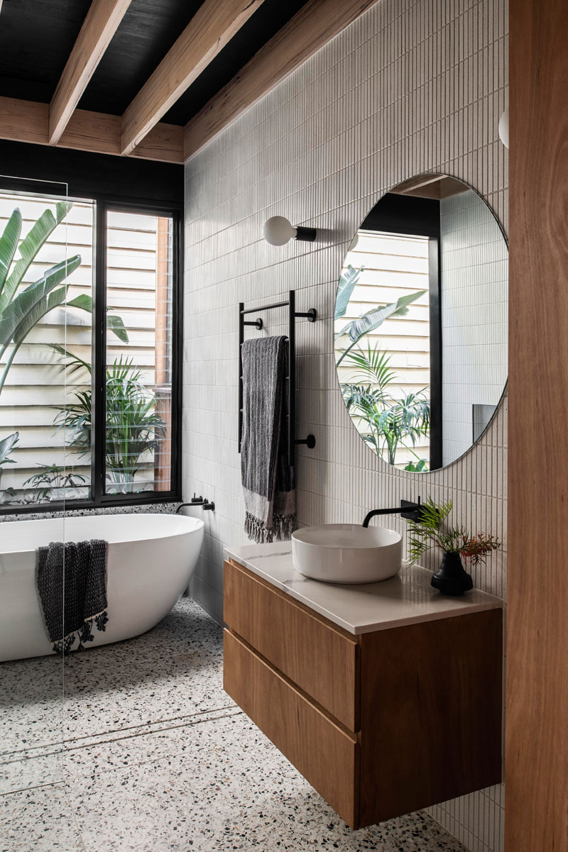 In the bathroom, tiles cover the walls, while a freestanding bathtub is positioned beneath the window, and a minimalist glass shower screen allows the natural light from the window to filter throughout the room. #ModernBathroom #BathroomDesign #TiledWalls #RoundMirror