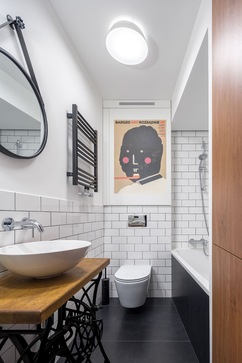 This modern bathroom features large black tiles on the floor, a show/bath combo, white subway tiles with dark grout, a hanging round mirror, and a custom vanity whose stand is made from the base of an old sewing machine. #ModernBathroom #BlackFloorTiles #WhiteSubwayTiles #BathroomDesign