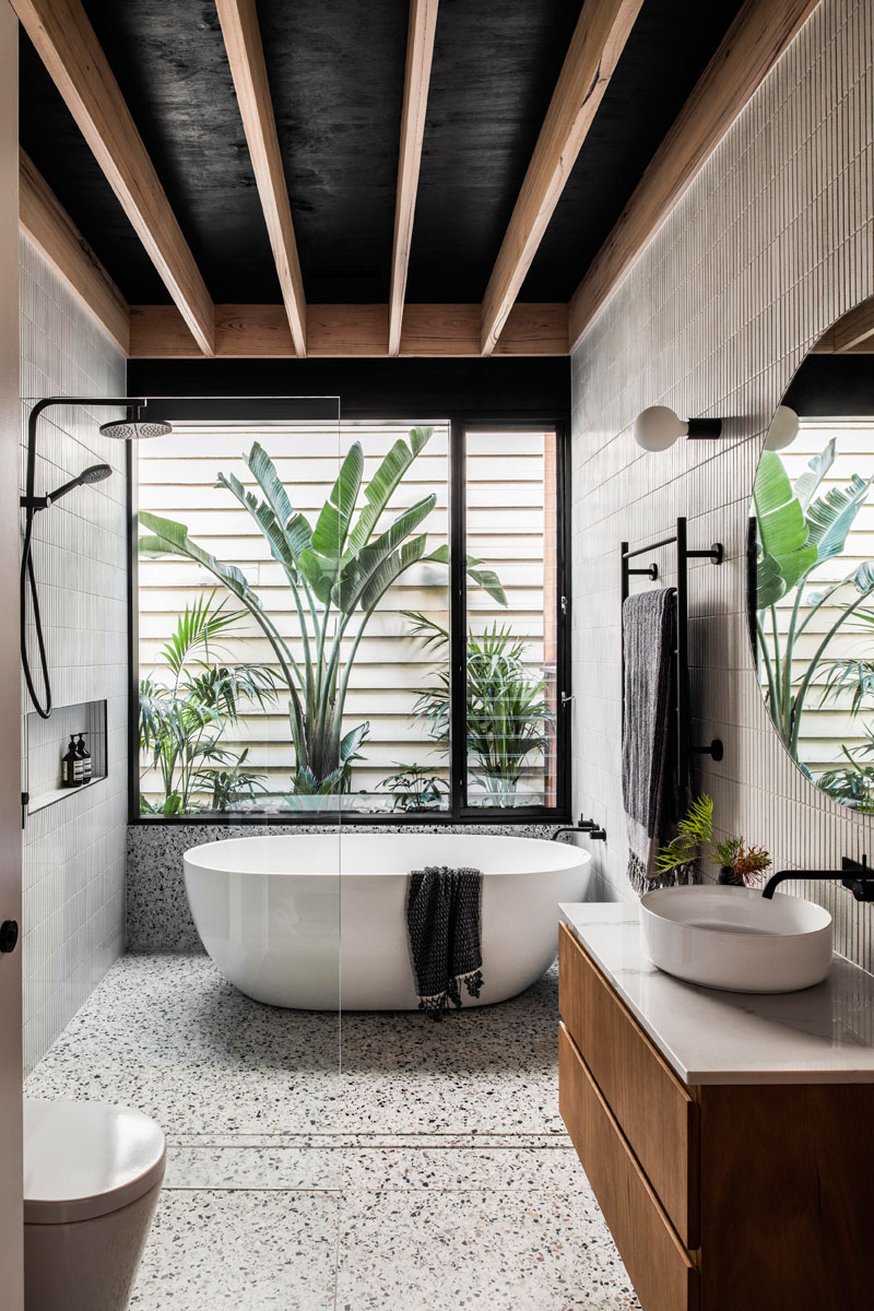 In the bathroom, tiles cover the walls, while a freestanding bathtub is positioned beneath the window, and a minimalist glass shower screen allows the natural light from the window to filter throughout the room. #ModernBathroom #BathroomDesign #TiledWalls