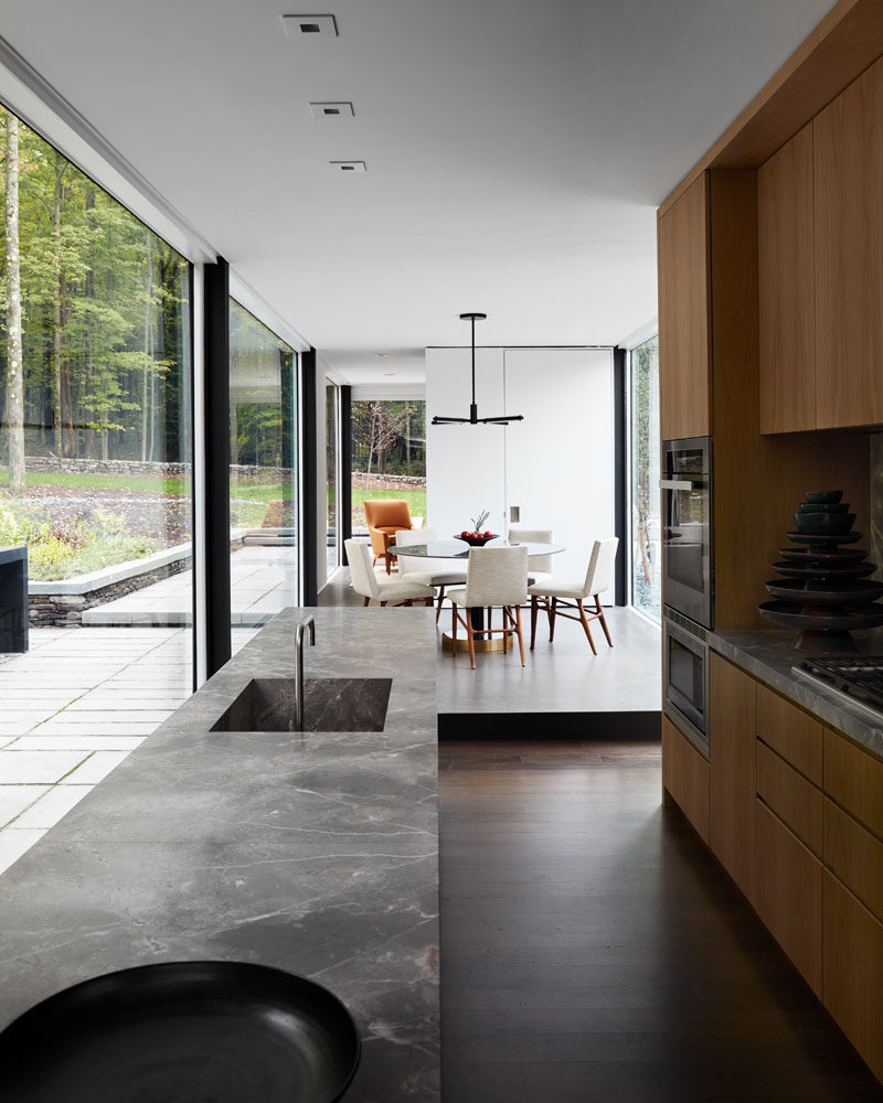 In this modern kitchen, wood cabinets have been combined with a grey countertop for a contemporary look, while floor-to-ceiling glass walls fill the room with natural light. #Kitchen #Windows #GlassWalls
