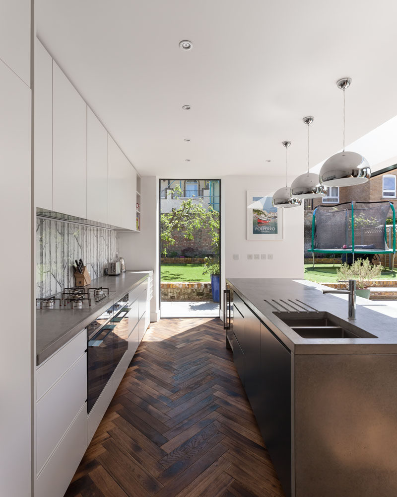 A nature-inspired backsplash complements the raw materiality of concrete countertops. Three mirror-polished pendant lights hang above the island, and help to create a modern industrial appearance. #ModernKitchen #StainedWoodFlooring #Window #ConcreteCountertops