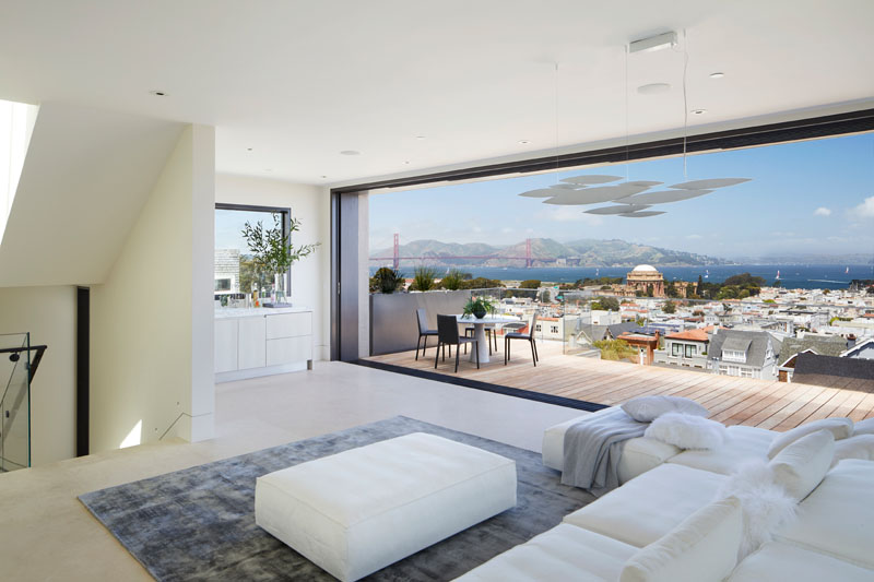 This modern house has a living room furnished in white and grey, that opens up to a balcony with city and water views. #LivingRoom #ModernInterior