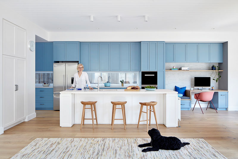 In this modern kitchen, matte light blue cabinets line the wall and transform into a small built-in home office with a bench, while a large white kitchen island provides additional seating. #LightBlueKitchen #ColorfulKitchen #KitchenIdeas #HomeOffice