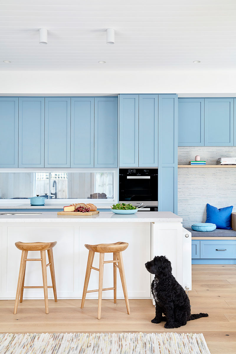 In this modern kitchen, matte light blue cabinets line the wall and transform into a small built-in home office with a bench, while a large white kitchen island provides additional seating. #LightBlueKitchen #ColorfulKitchen #KitchenIdeas #HomeOffice