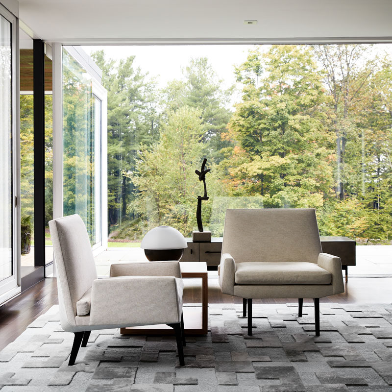 This modern sitting area has a textured grey rug, while floor-to-ceiling glass walls provide uninterrupted views of the trees outside. #SittingRoom #LivingRoom #InteriorDesign