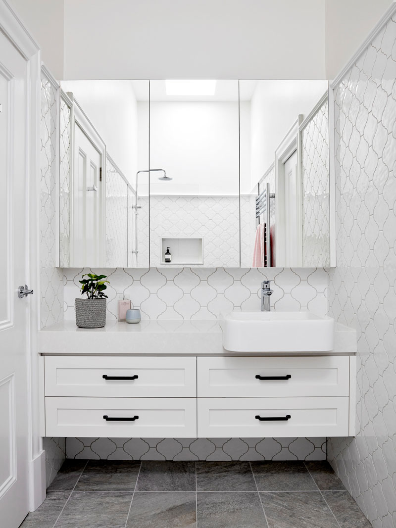 In this contemporary bathroom, an arabesque tile pattern in white covers the walls, while the wall-to-wall mirror reflects the shower and the natural light from the skylight. #WhiteBathroomIdeas #ContemporaryBathroom #TileIdeas