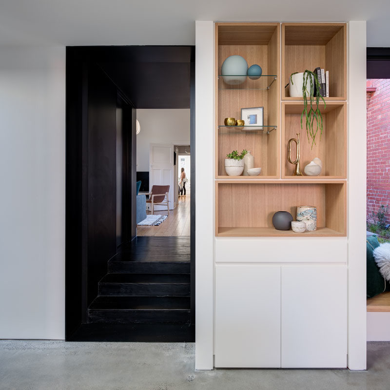 Shelving Ideas - In this modern home interior, the dark hallway opens up to the new addition that houses the main social areas. A built-in wood shelf is surrounded by white walls and cabinetry, a strong contrast to the black hallway. #ShelvingIdeas #WoodShelving #OpenPlanLiving