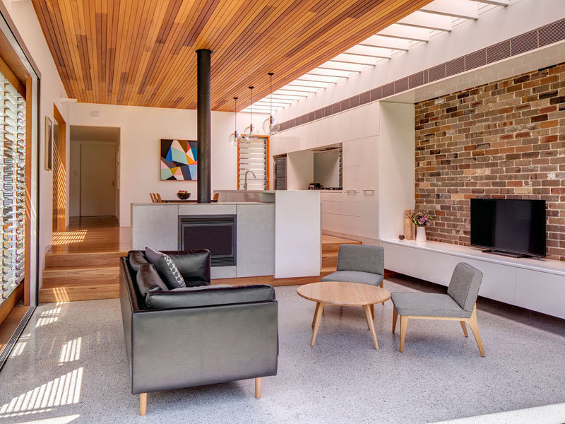 In this modern living room that features a time ceiling and recycled brick wall, there's fireplace and a few steps that provides separation between the living room and the dining room and kitchen. #LivingRoom #Fireplace #BrickWall #WoodCeiling