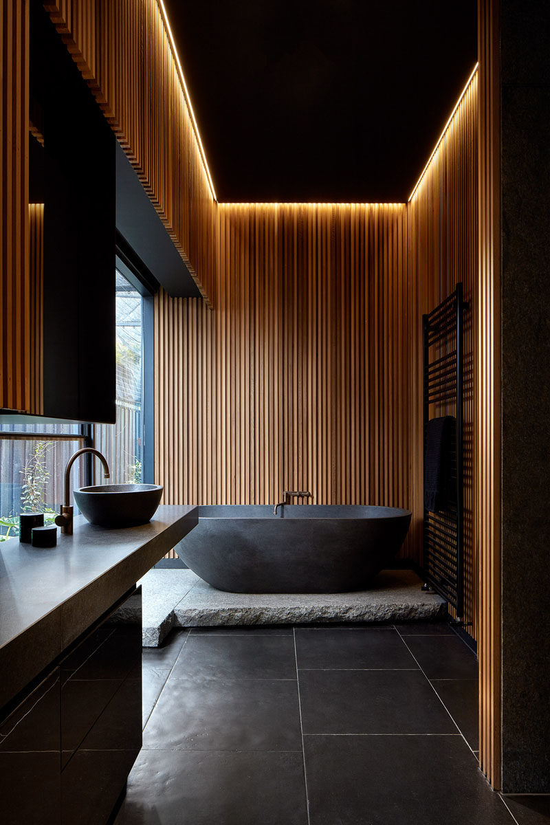 Bathroom Design Ideas - In this modern bathroom, hidden lighting creates a soft glow around the edges of the ceiling, while the wood slats create a backdrop for the black accents, like the bathtub, tile flooring, and window frame. #WoodSlats #BathroomDesign #ModernBathroom #WoodBathroom #BlackBathroom