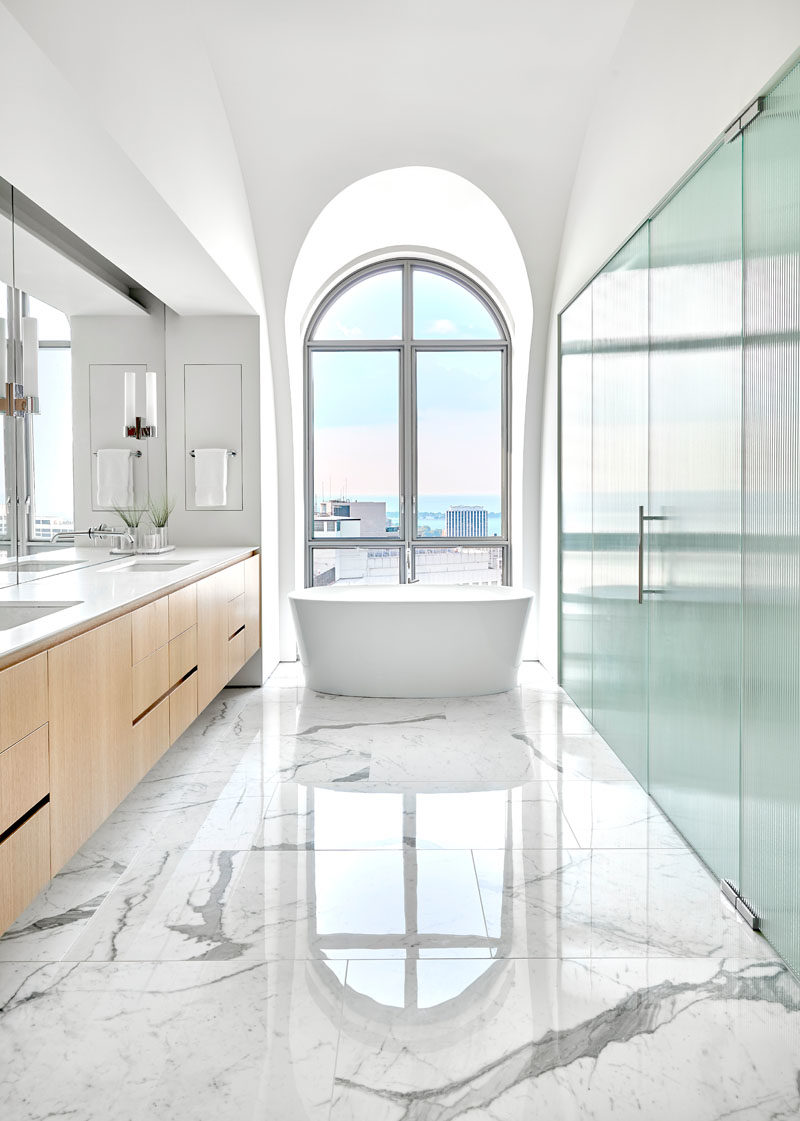 Bathroom Ideas - In this master bathroom, a freestanding bathtub sits directly in front of a large arched window, while a large vanity and mirror line the wall. #ModernBathroom #ArchedWindows #BathroomDesign