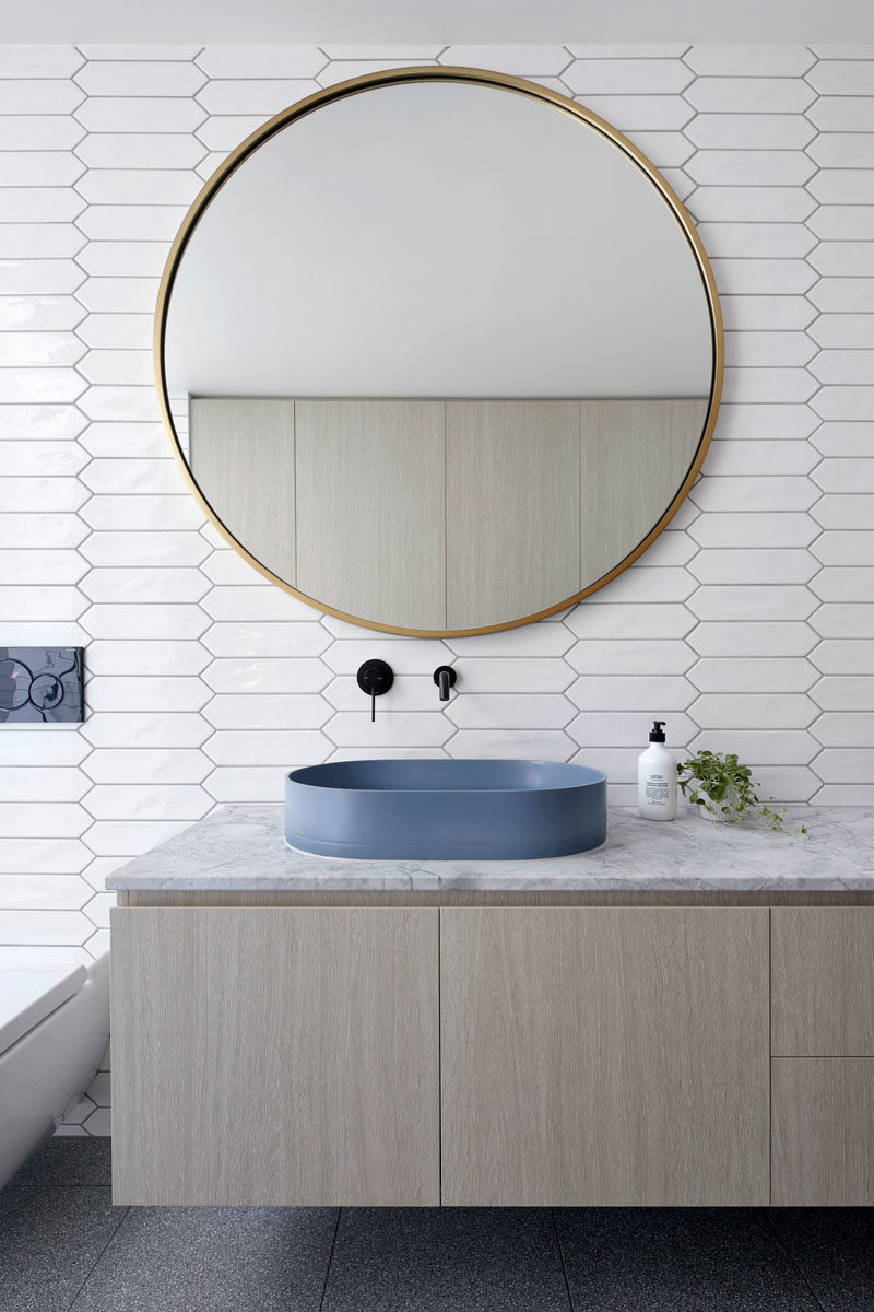 Bathroom Ideas - In this modern white bathroom,  picket tiles cover the walls, while the laundry is hidden away in wood cabinets opposite the vanity, and in the shower, there's a floor-to-ceiling window with views of a tiny garden at the side of the house. #BathroomIdeas #PicketTiles #WhiteTiles #HiddenLaundry #Windows