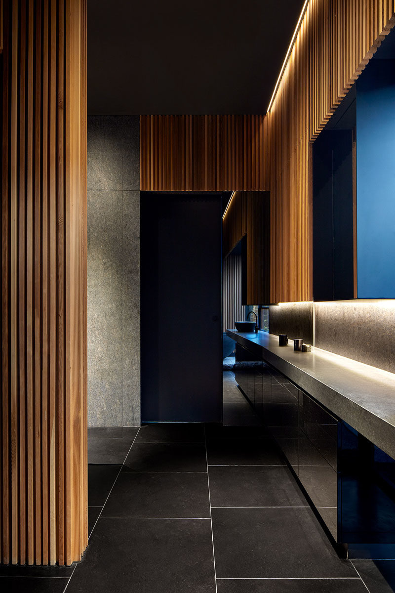 Bathroom Design Ideas - In this modern bathroom, hidden lighting creates a soft glow around the edges of the ceiling, while the wood slats create a backdrop for the black accents, like the bathtub, tile flooring, and window frame. #WoodSlats #BathroomDesign #ModernBathroom #WoodBathroom #BlackBathroom