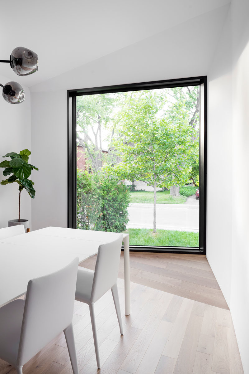 This modern dining room is furnished with a white dining table surrounded by white chairs, while a large picture window frames the street view. #DiningRoomIdeas #WindowIdeas #ModernDiningRoom #BlackFramedWindow