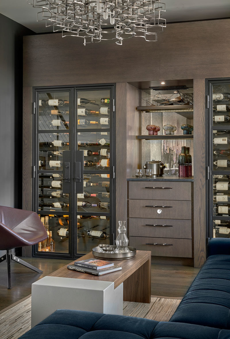 Wine Room Ideas - This modern wine room has been furnished with a ink blue sectional, has custom-built wine chillers, and a chandelier by Lucy Slivinsky, that's made of metal car parts. #WineRoom #WineStorage #Lounge #HomeBar