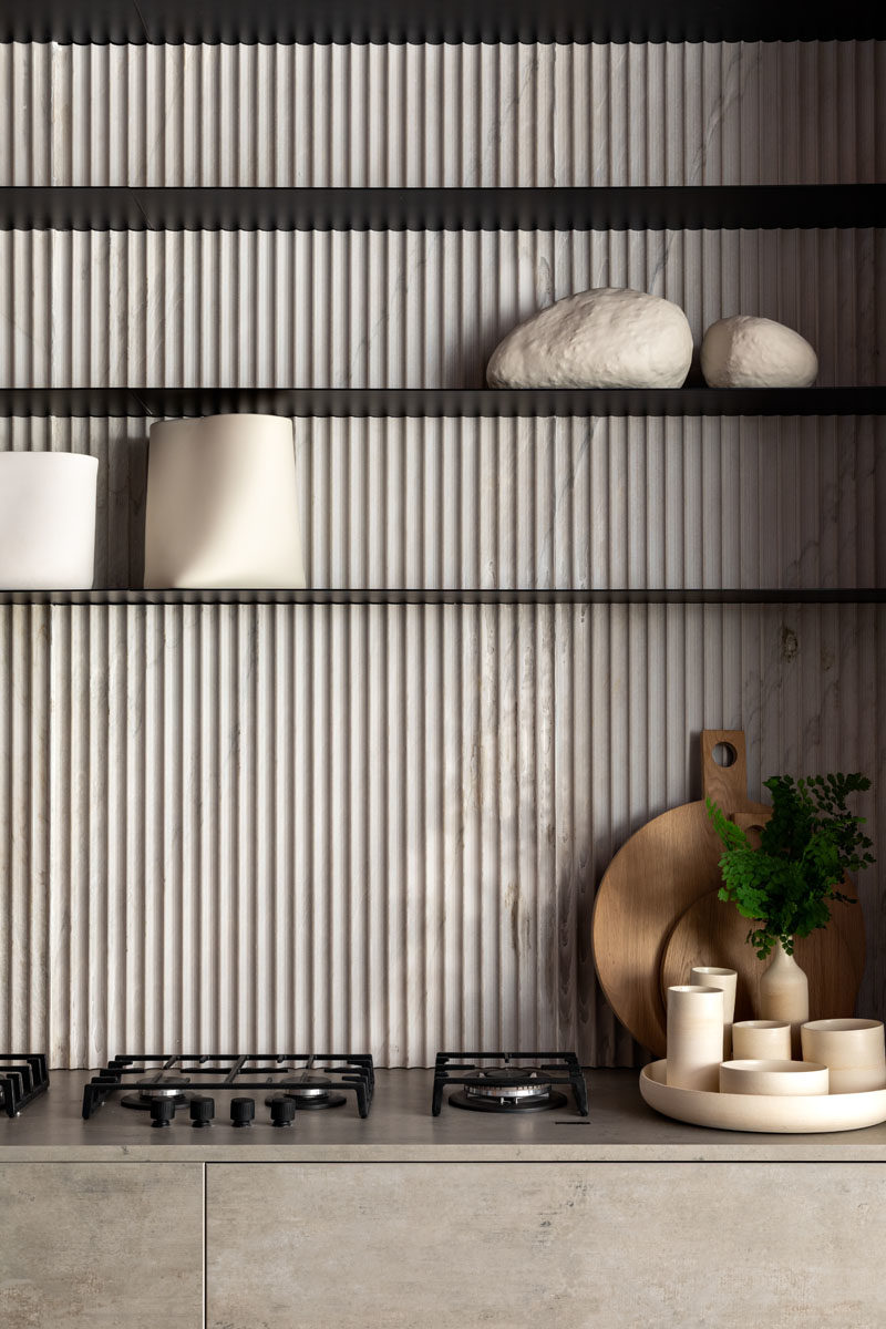 Kitchen Ideas - In this modern kitchen, a textured backsplash has been made from the same stone material as the countertops and cabinets. Thin minimalist black shelves provide a contrasting element, and complement the integrated burners. #KitchenIdeas #KitchenDesign #ModernKitchen