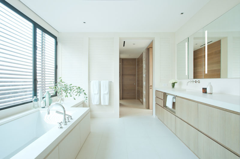 Bathroom Ideas - In this bathroom, a built-in bathtub sits below the window, while on the opposite wall, a wood vanity runs the length of the room and is topped with a light counter. #BathroomIdeas #ModernBathroom #WhiteBathroom
