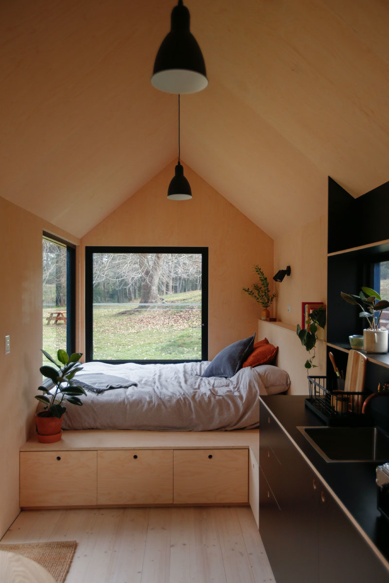 At one end of this modern tiny cabin is the open bedroom, that has two large windows, providing natural light and views of the trees outside. Storage has been added underneath the bed, and also acts as a bench to sit on. #TinyCabin #TinyHouse #TinyHome #Bedroom #Windows