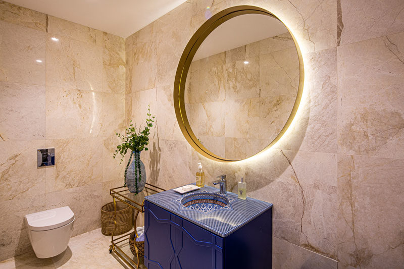 Bathroom Ideas - In this modern bathroom, a blue vanity has a decorative top, while the large round mirror is backlit, and a small side table adds a place to display a vase. #BathroomIdeas #ModernBathroom #BathroomDesign