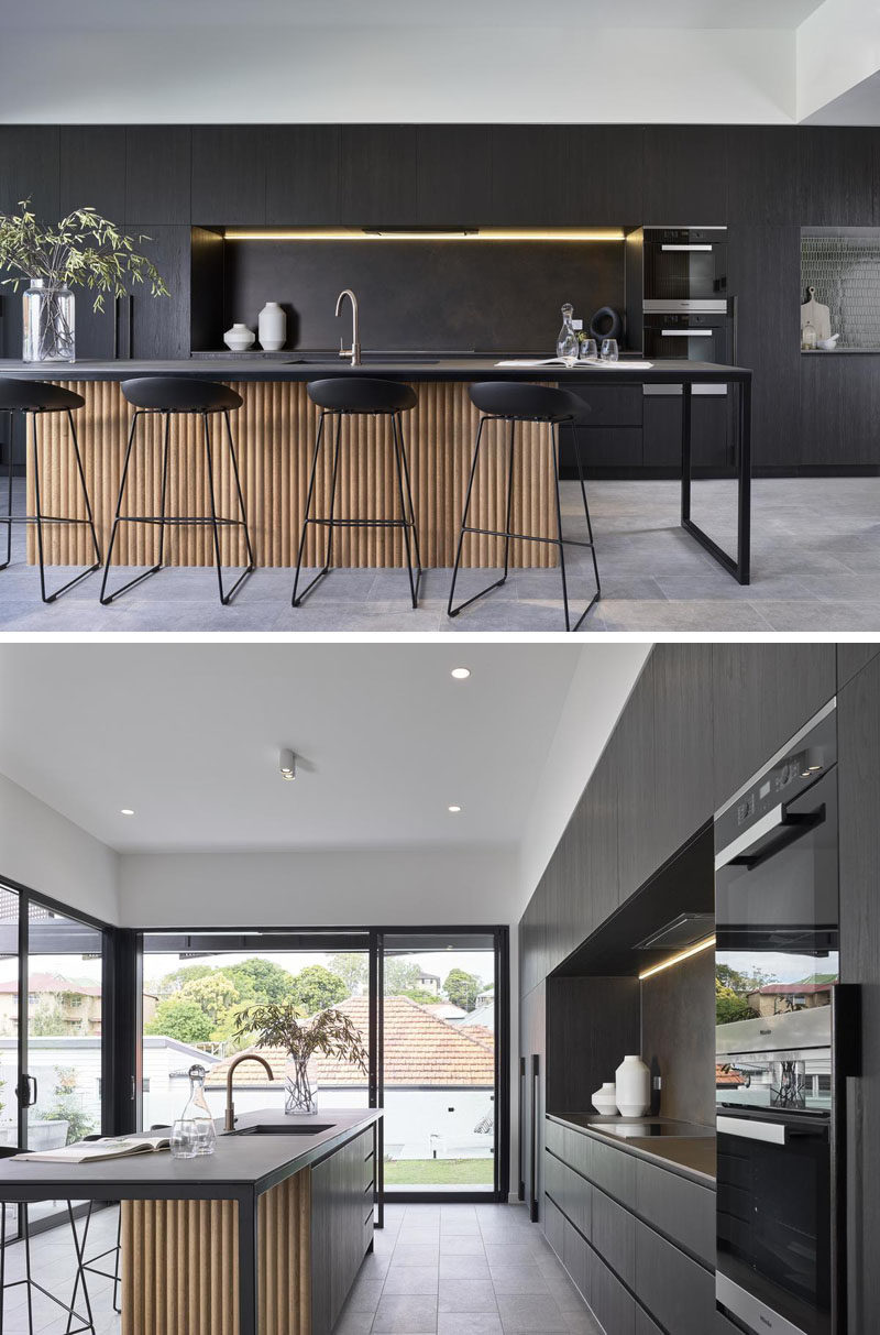 Kitchen Ideas - In this modern kitchen, minimalist dark cabinets line the wall, and an island with room for bar seating, features a wood accent. #KitchenIdeas #DarkKitchen #ModernKitchen #KitchenDesign