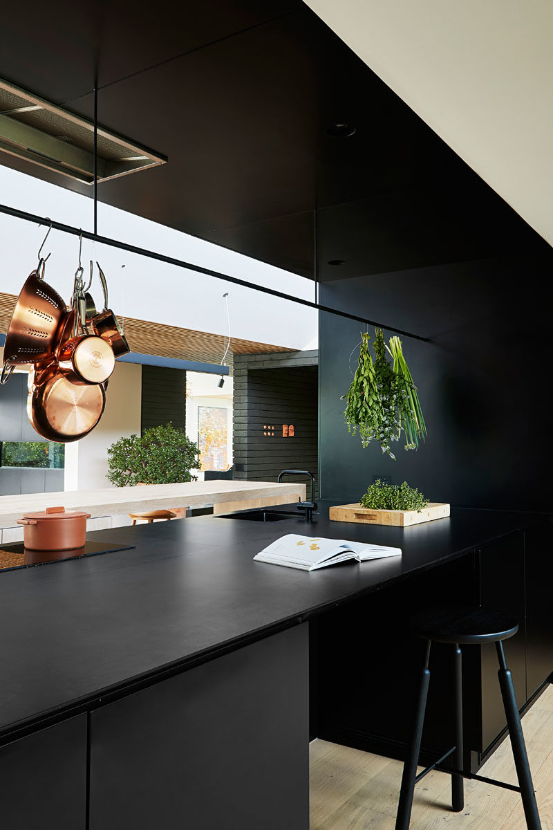 Kitchen Ideas - In this modern kitchen, minimalist black cabinets line the wall, and a large island provides ample counter space and a place for sitting. A void in the cabinets allows for connectivity between the kitchen and the pantry, that's located behind the cabinets. #KitchenIdeas #BlackKitchen #KitchenDesign #ModernKitchen