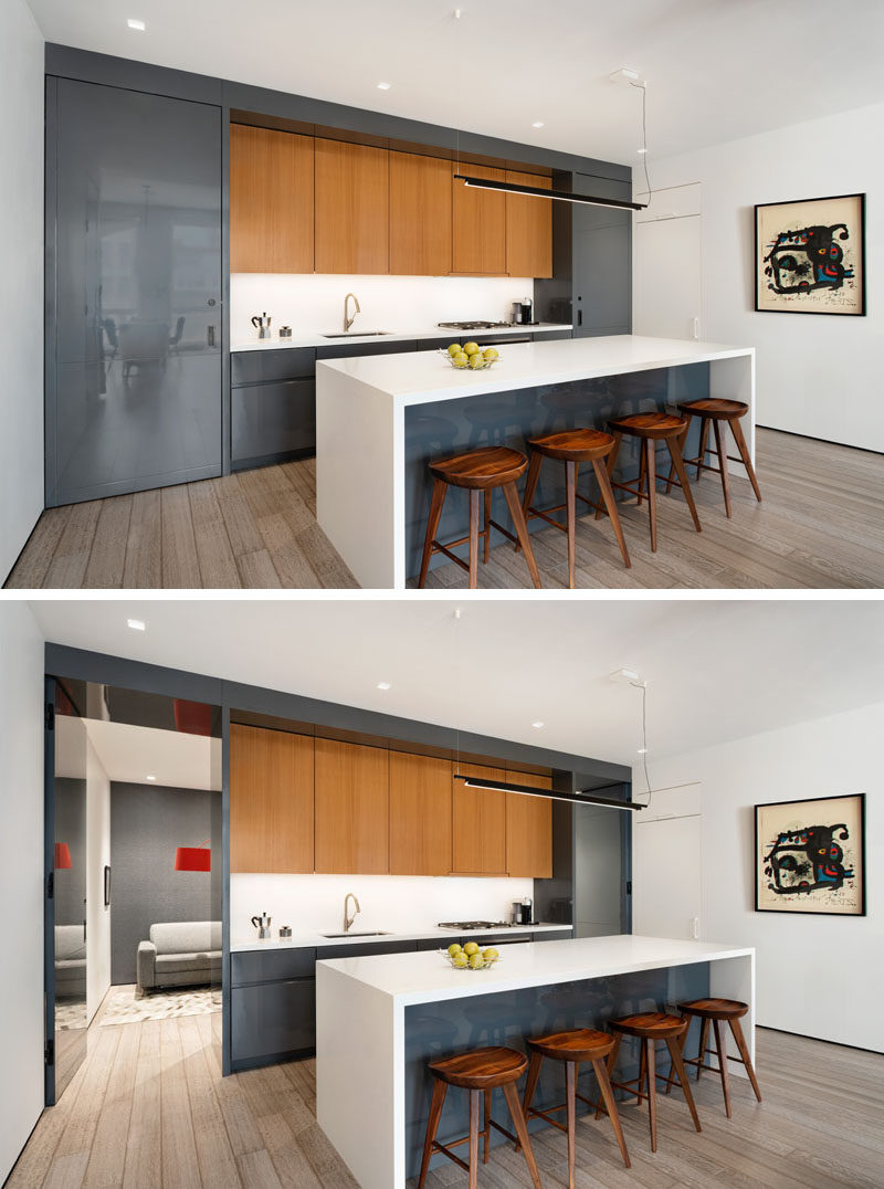 Kitchen Ideas - In this modern kitchen, high gloss gray lacquer cabinets have been used to complement the living room TV unit. The kitchen also features custom flush pivot doors on either end, built to look like part of the kitchen cabinetry. One of the doors reveals a hidden office, while the other leads to a guest bathroom. #KitchenIdeas #ModernKitchen #GreyCabinets #KitchenIsland