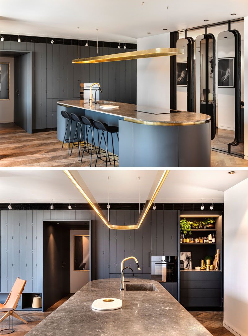 In this modern kitchen, there's a spacious island that's been designed with an oval shape. It includes touches of brass and is clad in curving cupboards that are made from wood and painted in a coal-grey with a blue tinted pigment. The island countertop is brown marble with light colored veins, and the light fitting above the island is also brass, pressed into an oval shape. #ModernKitchen #OvalIsland #KitchenIslandIdeas #KitchenIdeas #KitchenDesign