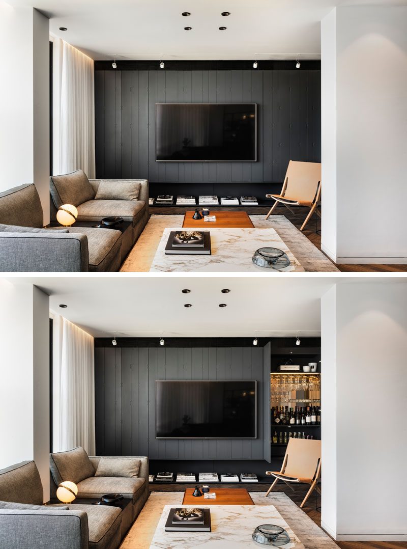 In this modern living room, there's a grey wall that runs the width of the apartment, and has hidden design features, like a bar with a mirrored wall and stemware storage. #LivingRoomIdeas #ModernLivingRoom #HiddenBar