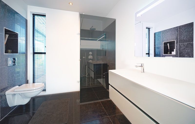 In this modern bathroom, a niche has been built into the wall above the toilet, while dark glass frames the shower, and a floating white vanity sits below a large mirror. #ModernBathroom #BathroomNiche #FloatingVanity #DarkGlass