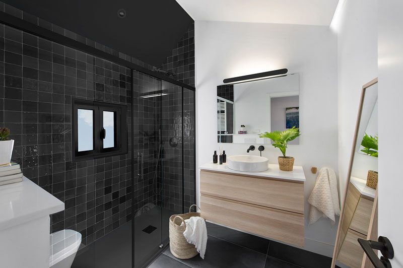 In this modern bathroom, square black tiles define the shower and allow the window frame to blend in, while a light wood vanity adds a natural touch to the room. #ModernBathroom #BlackShower #BlackTiles #BathroomIdeas #WoodVanity