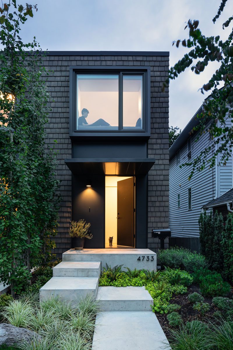 The path to the front door of this modern house runs alongside a line of trees that creates a barrier from the sloped driveway, and provides a view of one of the two bay windows on the second floor of the house. #ModernHouse #ModernBayWindow #DarkShingles #ModernLandscaping #Path