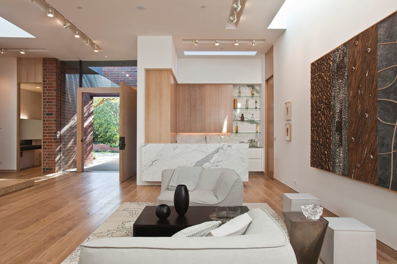 This modern house has an open living room with an adjacent bar that features wood cabinetry and stone materials. #ModernLivingRoom #HomeBar