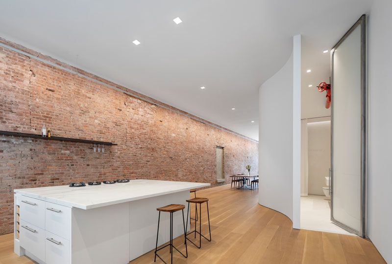 The old brick wall runs the full length of this modern loft, while on the opposite wall, a curved wall hides the bathroom behind a 13 foot tall custom sand-blasted pivot door. #ModernLoft #LoftIdeas #Apartment #BrickWall