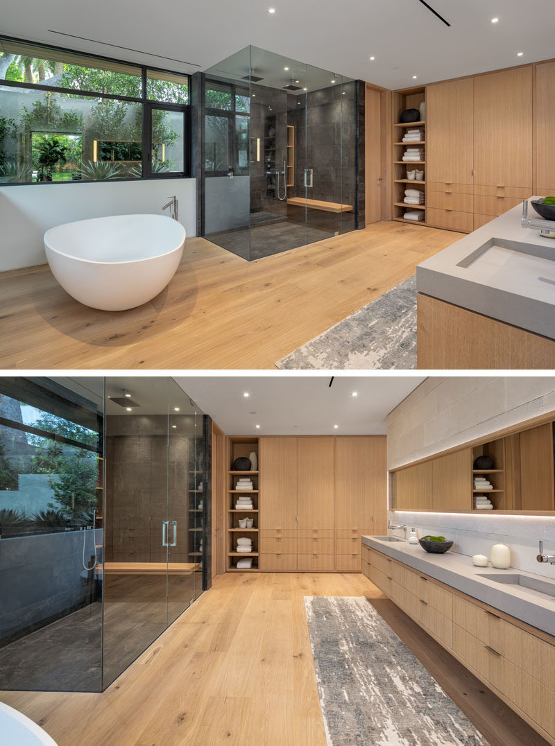 A modern en-suite bathroom with a sculptural freestanding white bathtub, a glass enclosed shower for two, windows with a view of the plants outside, and wood cabinetry. 