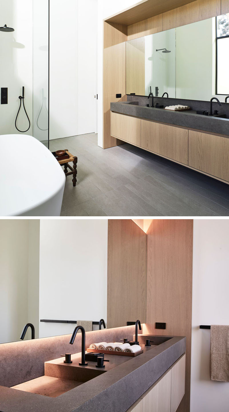 In this modern master bathroom, a wood and dark grey stone vanity sits below a large mirror with hidden lighting. #MasterBathroom #BathroomDesign #BathroomVanity