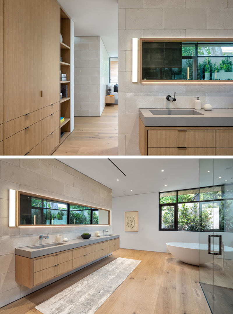 A modern bedroom has a long floating wood vanity that sits below a mirror with hidden lighting, and has a thick grey counter with built-in sinks.