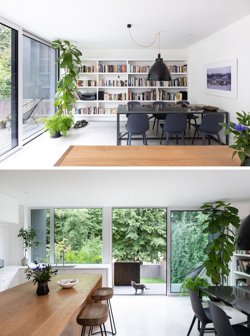 This modern open plan dining room has dark furnishings that contrast the bright white walls, while a shelving unit is filled with colorful books. A sliding glass door opens the room to the backyard where there's a deck and a lawn surrounded by trees. #DiningRoomIdeas #Bookshelf #SlidingGlassDoor