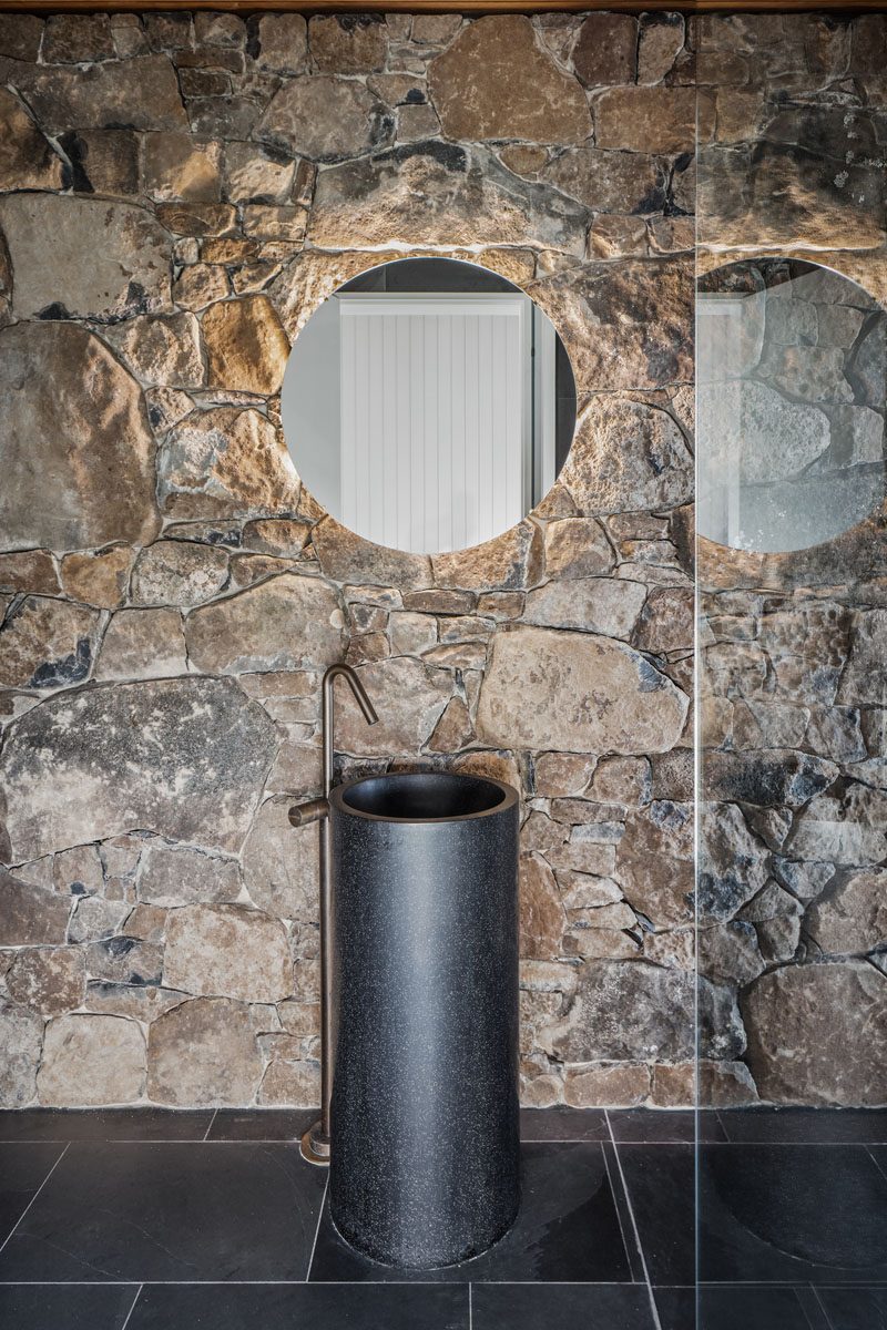 In this modern bathroom, the stone wall creates a backdrop for the minimalist black pedestal sink, while the backlit round mirror adds a soft glow to the space. #StoneWall #ModernBathroom #RoundMirror #BacklitMirror #BathroomIdeas