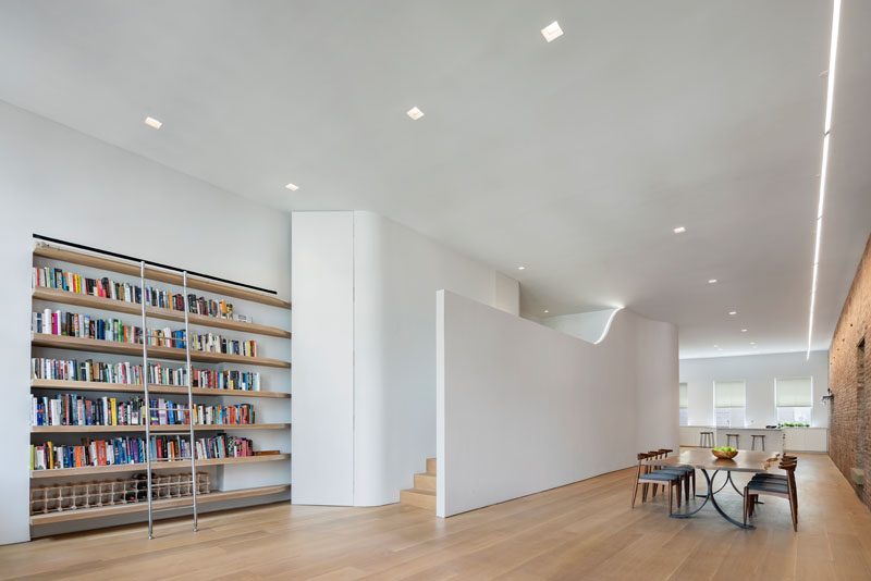 At the other end of this modern loft, there's a wall of solid oak library shelves near some stairs that lead up to a mezzanine that features a LED Light that lines the open wall. #ModernShelves #LoftApartment #ApartmentDesign