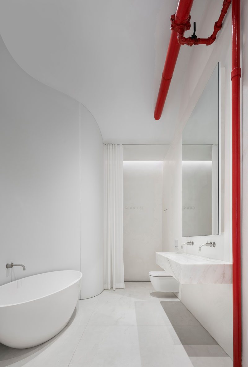 This modern bathroom includes ten foot long floor slabs of pale porcelain tile, white Imperial Danby stone for a cantilevered sink, and waterproof plaster walls. The old sprinkler main valve adds a bright pop of color to the white interior, while concealed rollers enable a large mirrored door to slide smoothly to the side, revealing a large cabinet with a soft LED light. #BathroomDesign #MinimalistBathroom #BathroomIdeas #WhiteBathroom