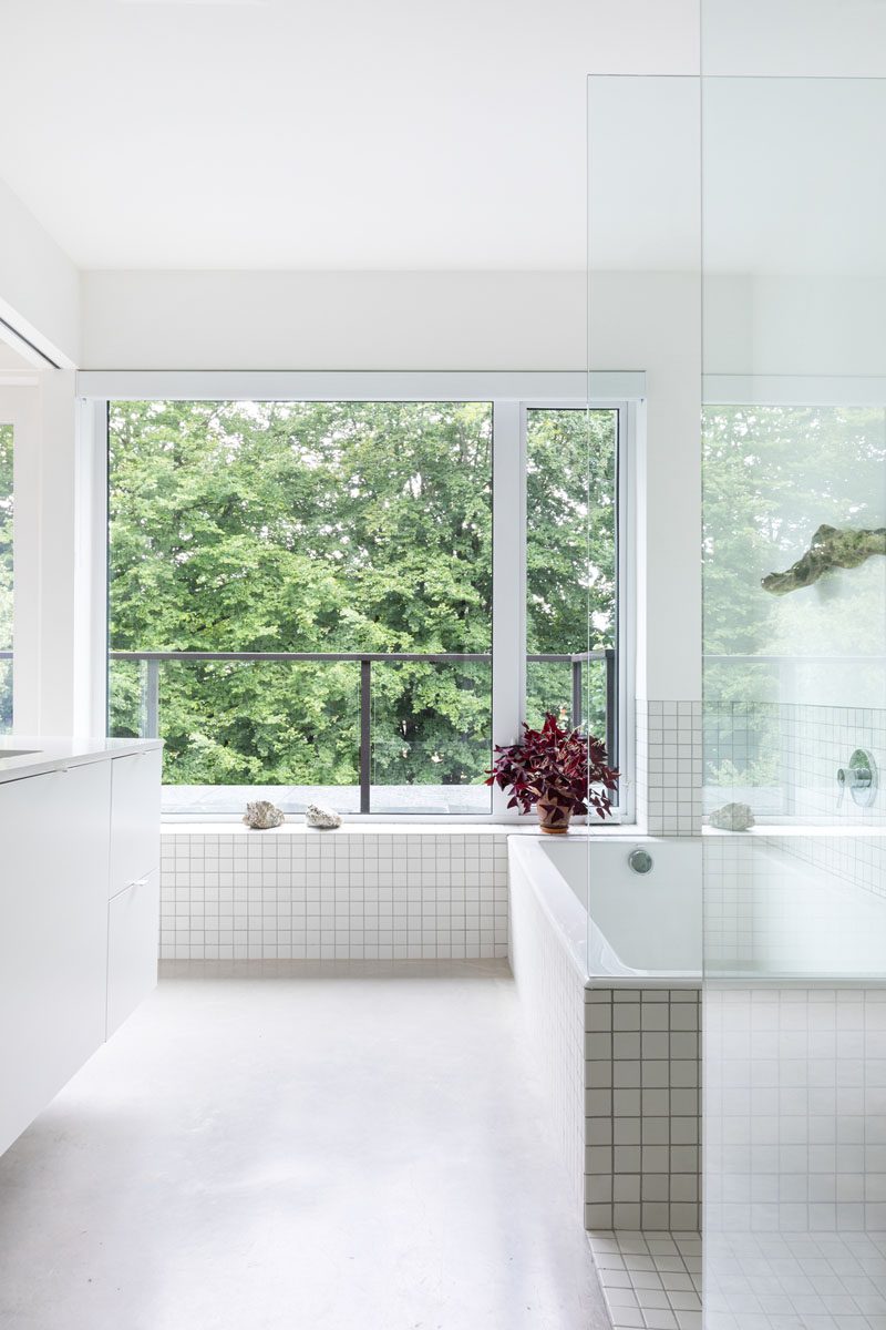 In this modern bathroom, large windows look out to the trees, while a built-in bathtub is surrounded by white square tiles, and is adjacent to a glass enclosed shower. #ModenBathroom #WhiteBathroom #BuiltInBathtub #ConcreteFloors