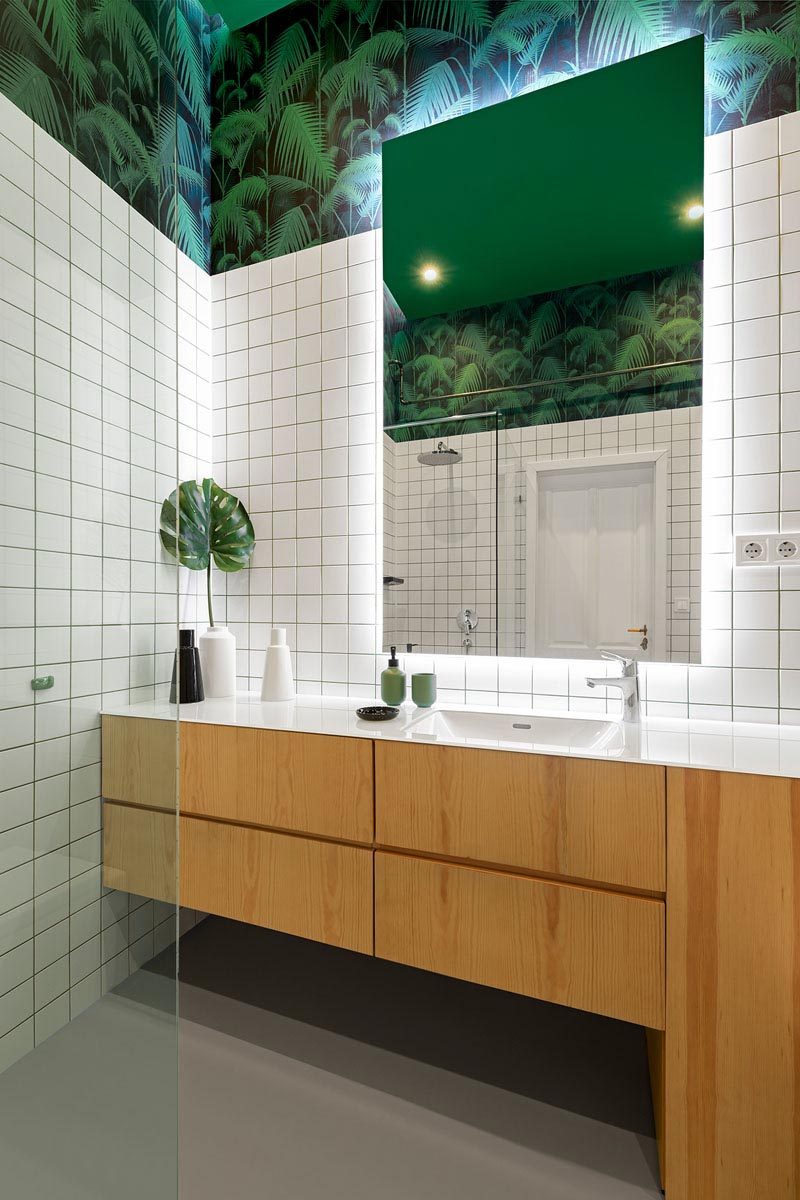 This modern bathroom has been decorated with square white tiles, a wood vanity, decorative wall coverings in a palm tree pattern, and a mirror with back-lighting. #Bathroom #ModernBathroom #GreenWhiteBathroom