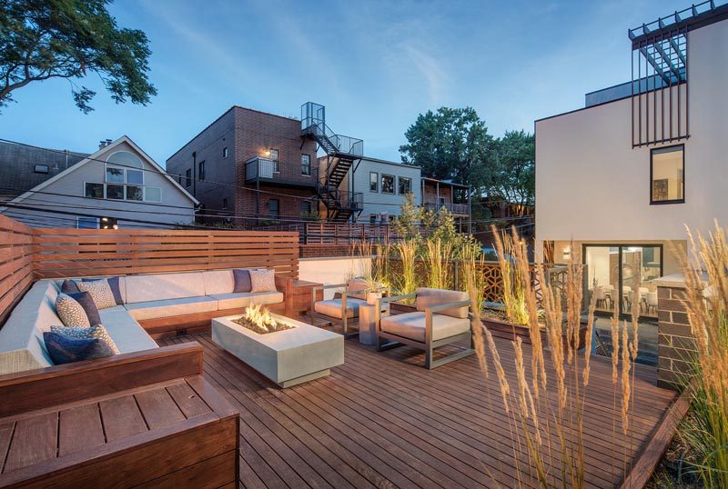 This rooftop deck has been furnished with a built-in corner sofa that has a small privacy screen, a pair of armchairs, a fire table, and tall plants. #RooftopDeck #Landscaping #Architecture #OutdoorSpace