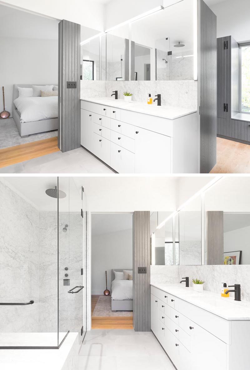 A minimalist white vanity with dual sinks and black hardware, sits below a mirror that wraps around the corner and is highlighted with accent lighting. #BathroomDesign #ModernBathroom #BathroomVanity