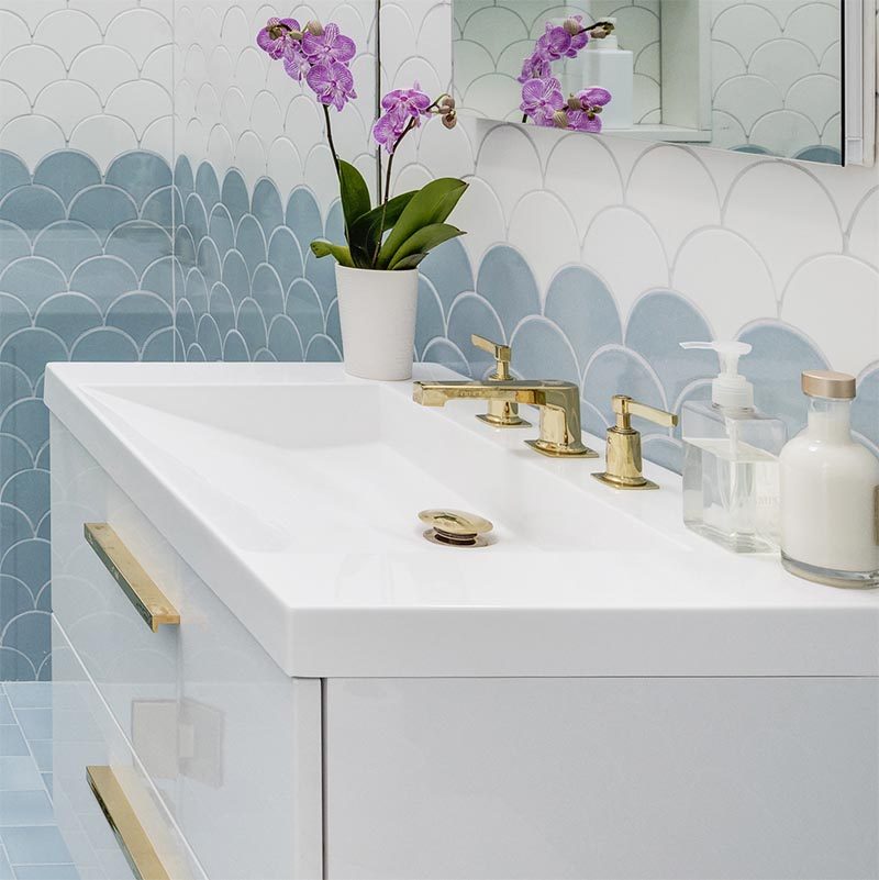 This modern bathroom is furnished with white vanity that has clean lines forming a rectangular shape, a built-in sloped sink, gold hardware in the form of the faucet, taps, drain, and drawer pulls.  #ModernBathroom #WhiteBathroomVanity #ModernVanity #BuiltInSink #GoldBathroomAccents