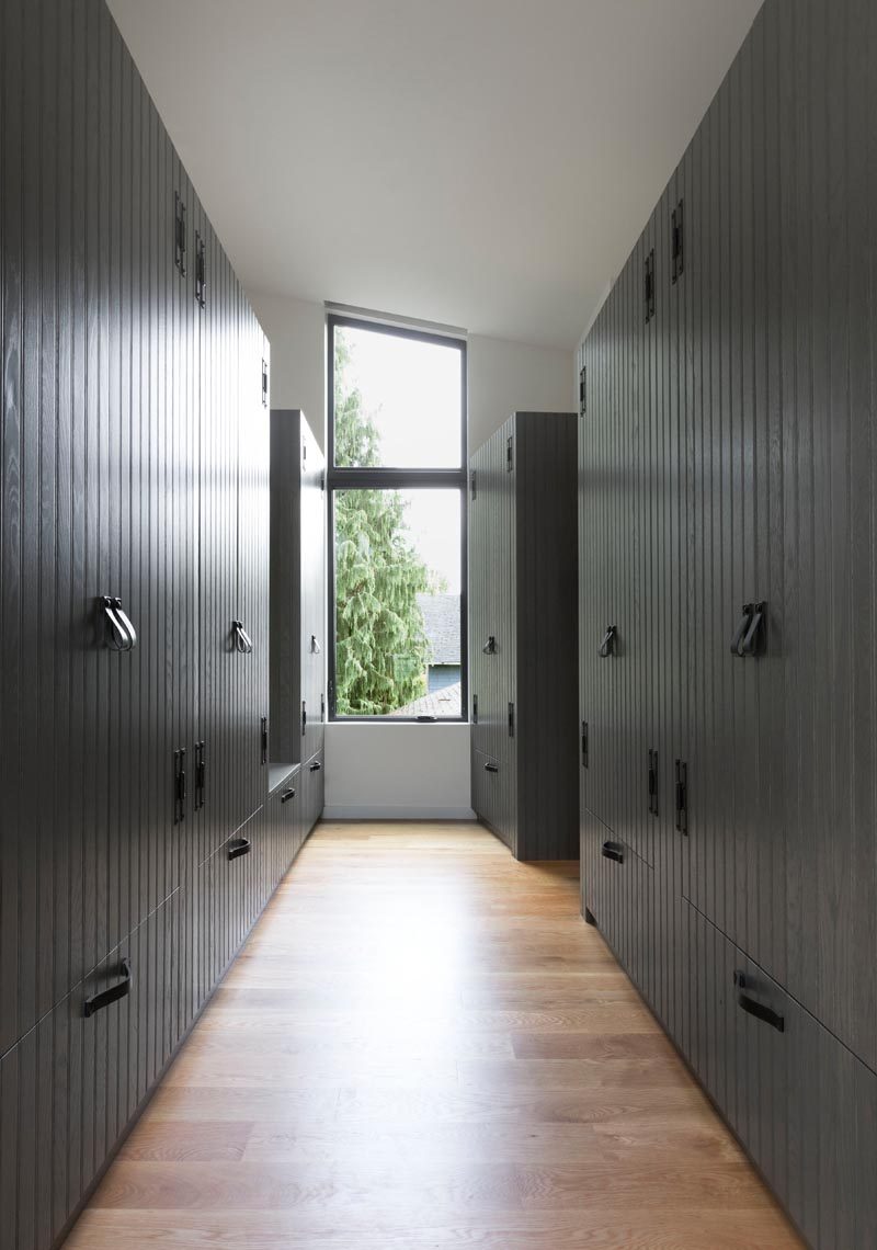 This modern walk-through closet has custom wood cabinetry with a dark gray wood paneling finish. A tall window at the end of the closet keeps the space bright, while a gap in the closet on the left hand side acts as a small bench or place to put your outfit when getting ready. #WalkThroughCloset #WalkThroughWardrobe #MasterSuite #Closets #Cabinets #DarkGreyCabinets