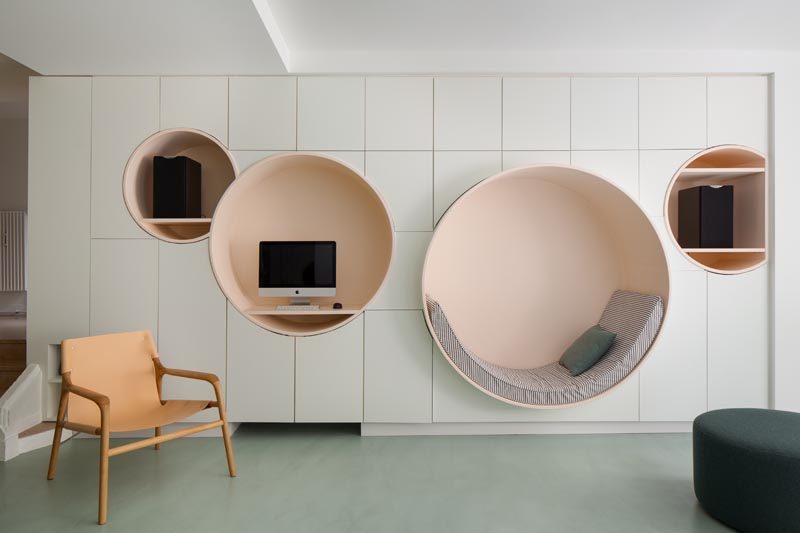 This modern living room has a wall of minimalist white cabinetry with multiple circular niches designed with different uses in mind. The largest circle has been defined as a reading nook, with an upholstered cushion that lines the bottom half of the shape, while the others have shelving for a computer and speakers. #ReadingNook #SeatingNiche #InteriorDesign