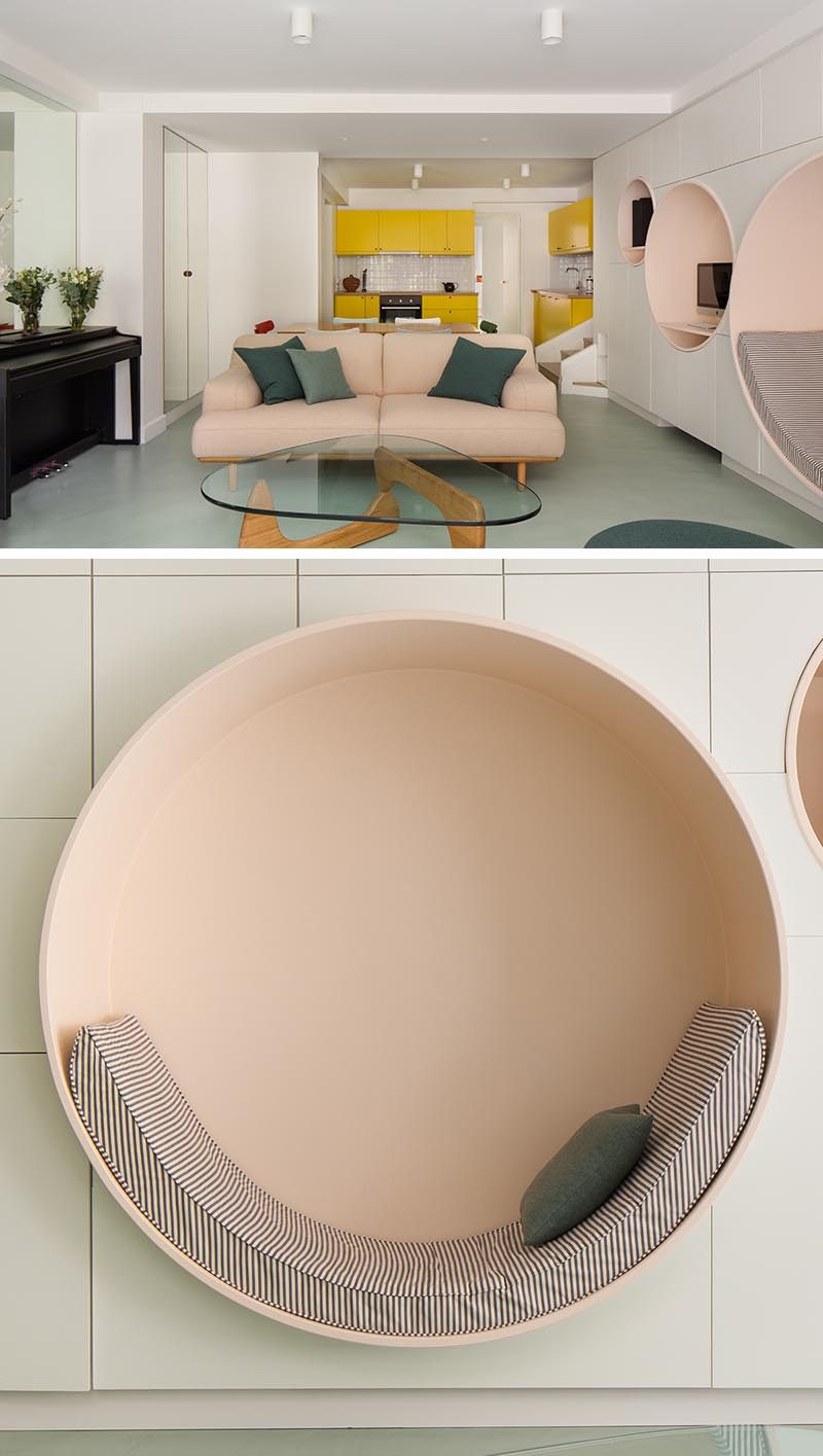 This modern living room has a wall of minimalist white cabinetry with multiple circular niches designed with different uses in mind. The largest circle has been defined as a reading nook, with an upholstered cushion that lines the bottom half of the shape, while the others have shelving for a computer and speakers. #ReadingNook #SeatingNiche #InteriorDesign