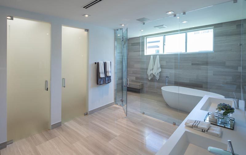 This modern master bathroom features a floating double-sink vanity, wood-like tile floors and walls, frosted doors, and a glass enclosed wet room that houses the shower and bath. #ModernBathroom #WetRoom #BathroomDesign #InteriorDesign #MasterBathroom